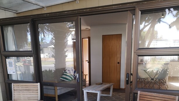 Un área de estar al aire libre con techo, sillas y banca de madera, un acceso por puerta de vidrio y un felpudo.