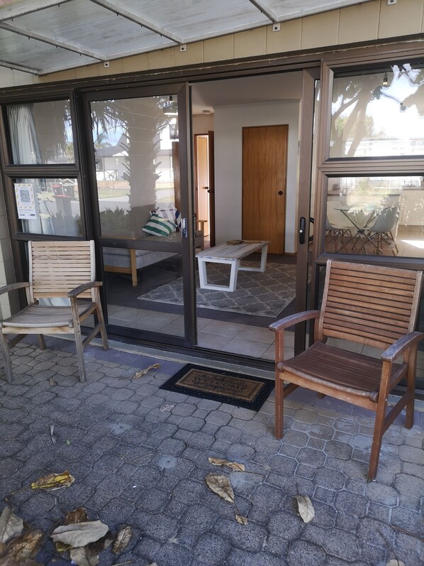 Un área de estar al aire libre con techo, sillas y banca de madera, un acceso por puerta de vidrio y un felpudo.