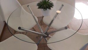 Una mesa de vidrio redonda con cuatro sillas blancas, una planta en maceta y una cocina al fondo.