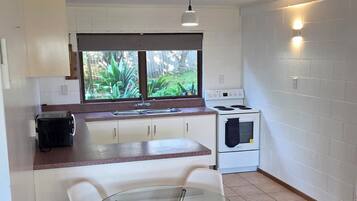 A compact kitchen with a glass table and chairs, a stove, a microwave, and a window with a view of greenery.