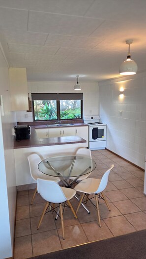 A compact kitchen with a glass table and chairs, a stove, a microwave, and a window with a view of greenery. - Just Beachy on Grant Place Papamoa Beach Pad (Papamoa)