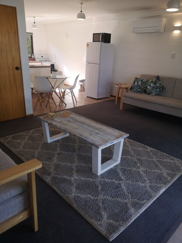 A living room with a wooden coffee table, a grey rug, a sofa with tropical patterned cushions, a white refrigerator, and a kitchen area with a microwave and a dining table with chairs.