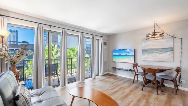 A modern living room with a sofa, a coffee table, a dining area with chairs, a TV mounted on the wall, and a balcony with curtains.