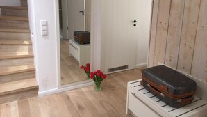 A room with a wooden floor, a white door, a white wall, and a wooden panel wall. There is a suitcase on a white cabinet and a vase with red flowers on the floor.