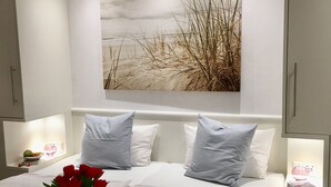 A modern bedroom with a large bed, white bedding, and a beach-themed wall art. There are two bedside tables with lamps and a tray with towels and red roses.