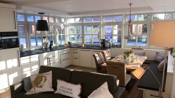 A modern kitchen with large windows, a dining area with a table and chairs, and a cozy seating area with a sofa and cushions.