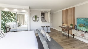 Habitación de hotel moderna con una cama grande, un escritorio con dos taburetes, un televisor y vistas a la cocina y al balcón.