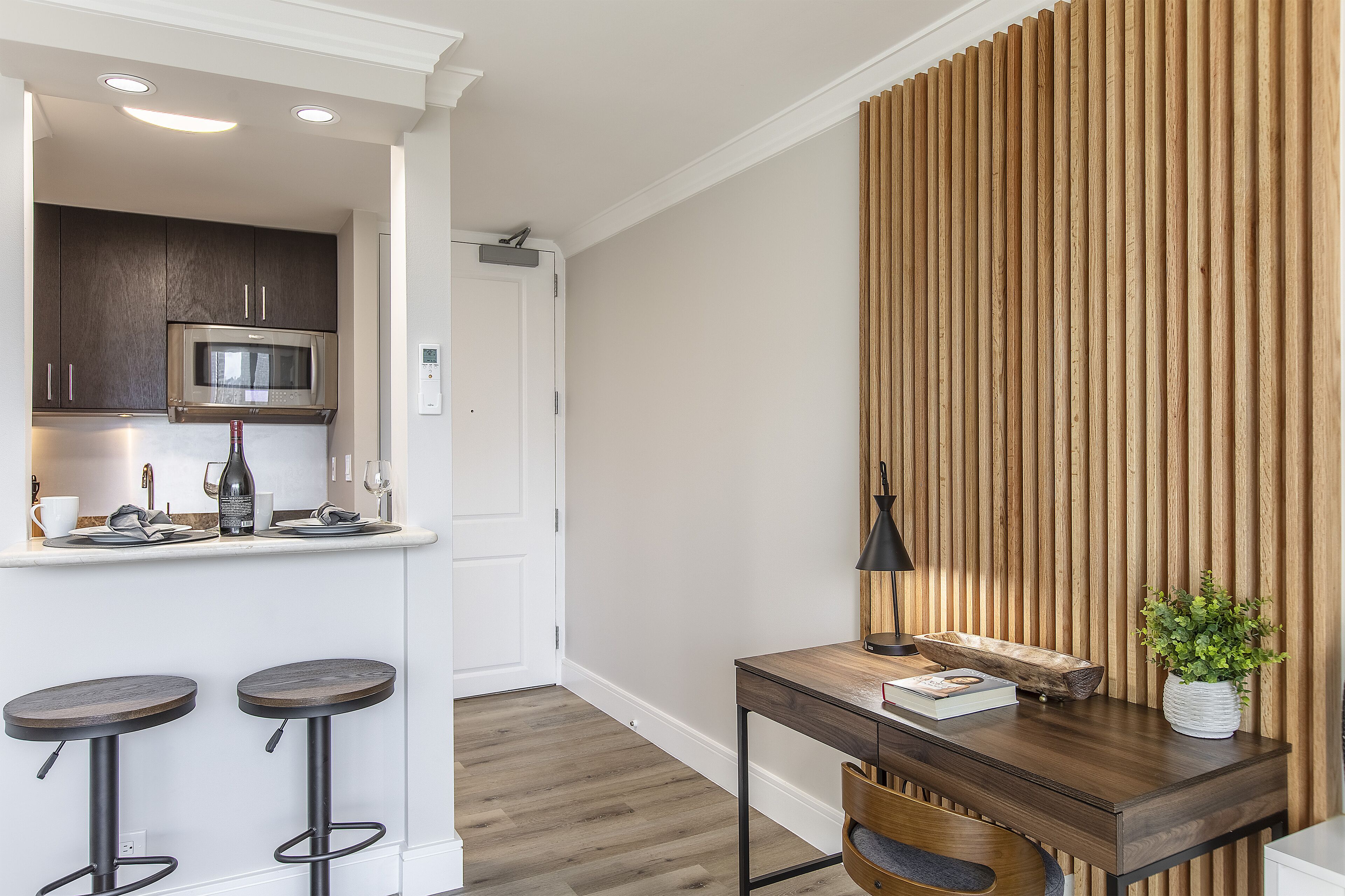A modern kitchen with dark cabinets, a stainless steel microwave, and a white countertop. There are two stools and a wooden table with a plant.