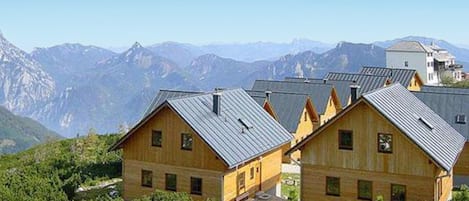 A row of wooden cabins with metal roofs, set against a backdrop of mountains and clear skies.