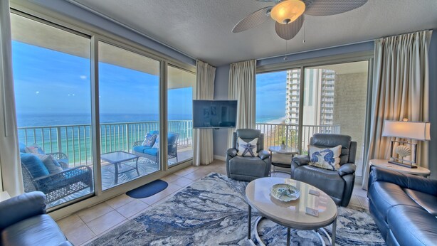 A spacious living room with a large balcony, a ceiling fan, a flat-screen TV, and a view of the ocean.