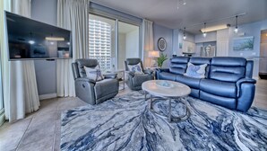 A modern living room with a flat-screen TV, grey leather sofas, a marble-patterned rug, and a ceiling fan.