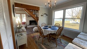 Comedor con una mesa redonda, sillas y una araña. Una escalera conduce a otra habitación, y se aprecia vista al exterior a través de amplios ventanales.