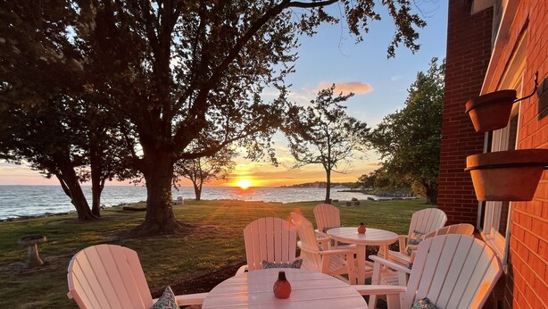 Un patio con sillas Adirondack y una mesa, con vistas a la puesta de sol sobre el agua.