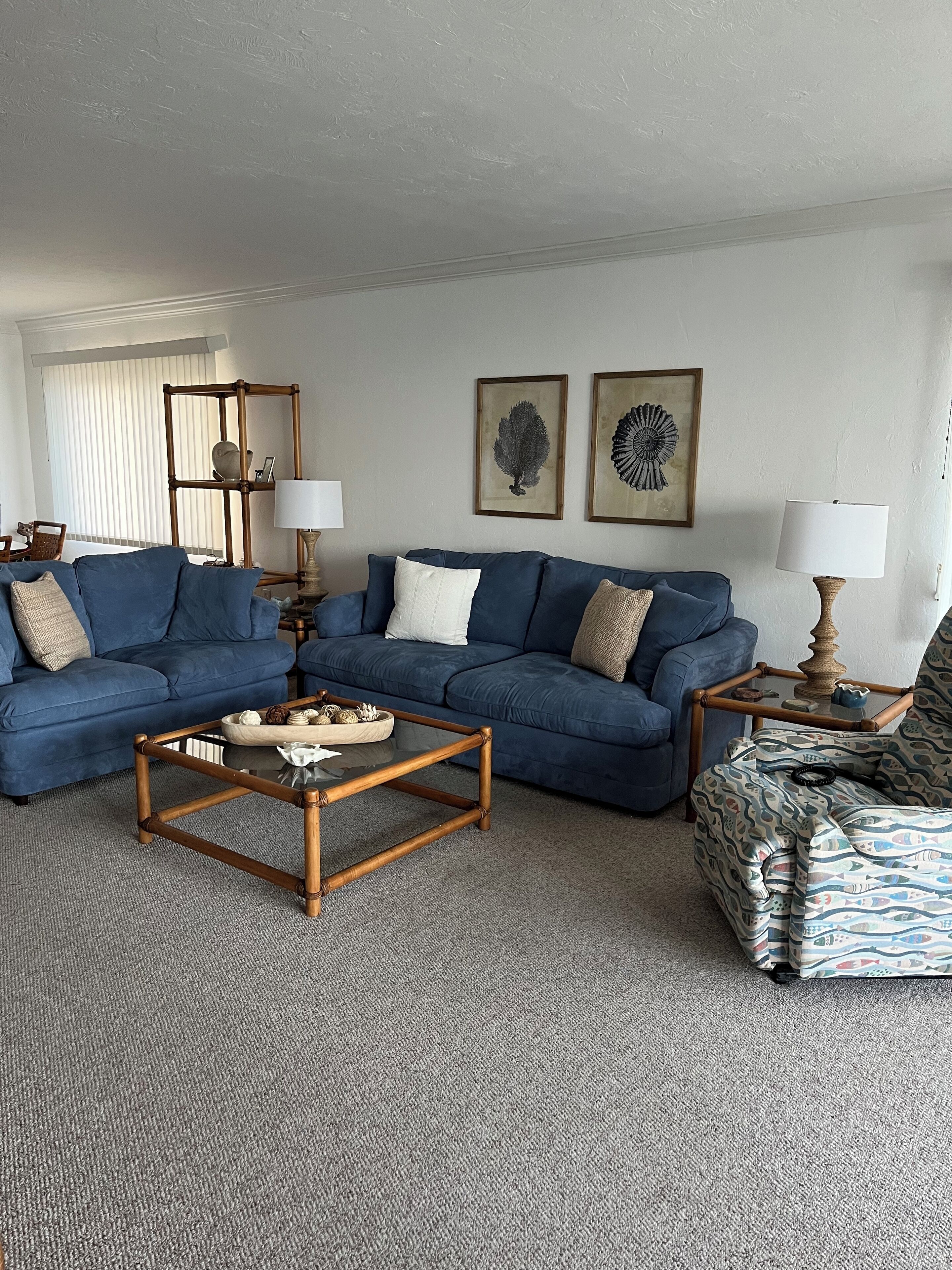 A living room with a blue sofa set, a wooden coffee table, and a patterned ottoman. There are two wall-mounted lamps and framed artwork above the sofa.