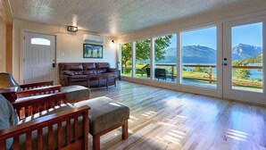 A spacious living room with a large window overlooking a lake and mountains, a leather sofa, wooden chairs, and a wooden floor.