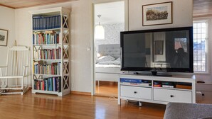 A living room with a large flat-screen TV, a bookshelf, a chair, and a bed with a window view. - Lovely home in Hakenäset with sauna (Hakenäset)