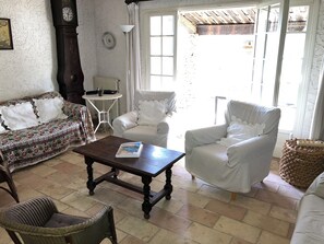 Uma sala de estar com um sofá estampado, uma poltrona branca, uma mesa de centro de madeira e uma cadeira de vime.