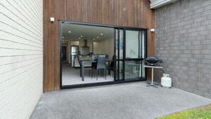 A modern patio with a glass door leading to an indoor dining area, a barbecue grill, and a propane tank. - Modern work-ready Hamilton home with sauna and EV charging (Waikato)