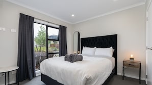 A modern bedroom with a large bed, a bedside table, a small table, and a window with curtains. - Modern work-ready Hamilton home with sauna and EV charging (Waikato)