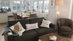 Un salon confortable avec un canapé, une table basse et un fauteuil. Il y a également un coin repas avec une table et des chaises. La pièce est dotée de grandes fenêtres et offre une vue sur l’extérieur.