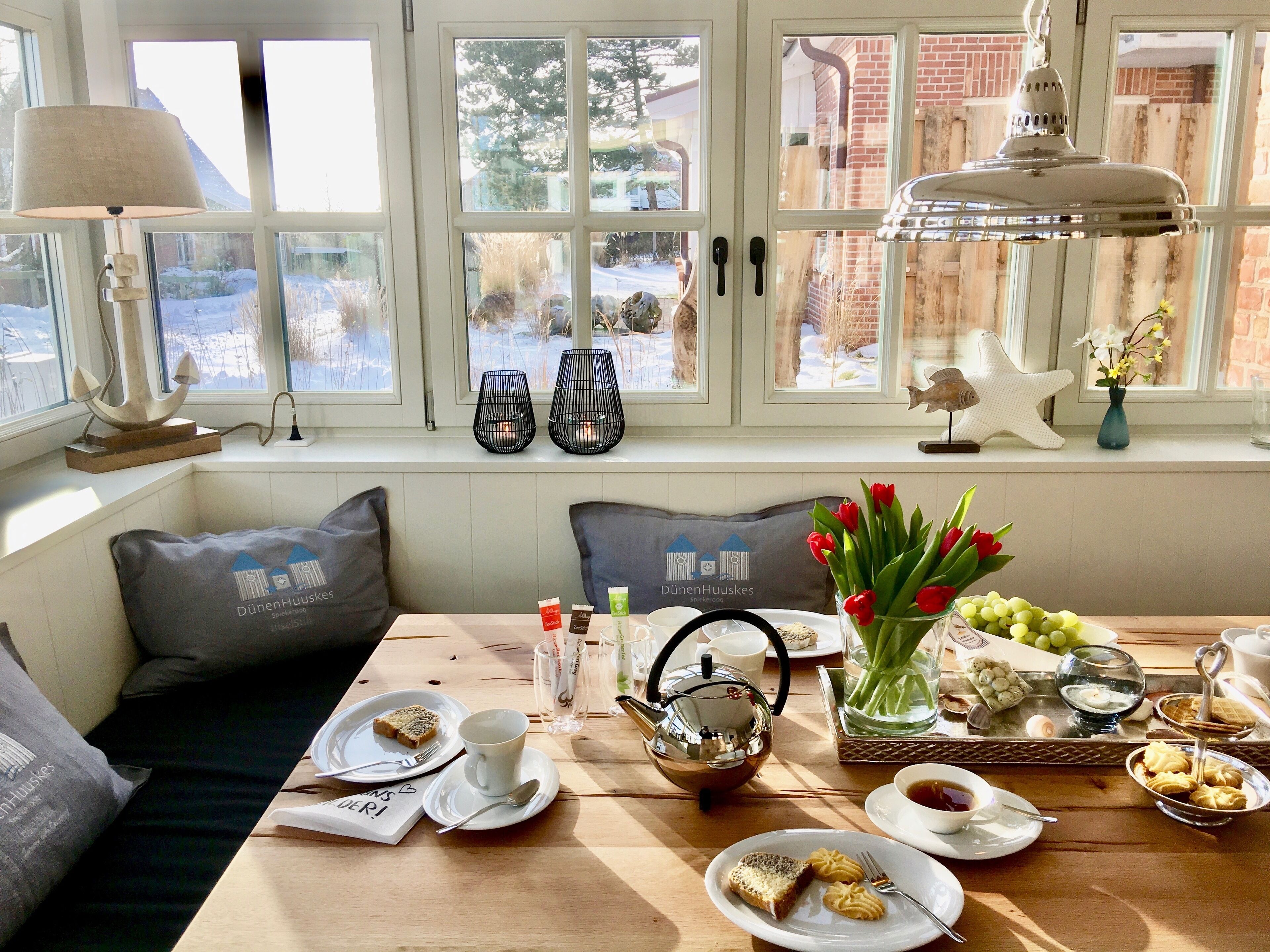 A dining area with a table set for two, featuring a variety of dishes, a teapot, and a vase with flowers.
