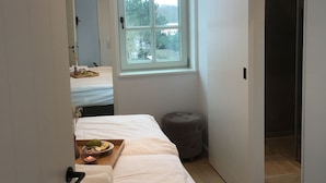Une chambre à coucher comprenant un lit, une fenêtre, une table de chevet avec un plateau, et une porte donnant sur une autre pièce.