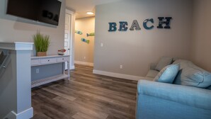 Una sala de estar con una televisión de pantalla plana, un estante montado en la pared con una planta, un sofá azul y una pared con la palabra 'BEACH' escrita en letras.
