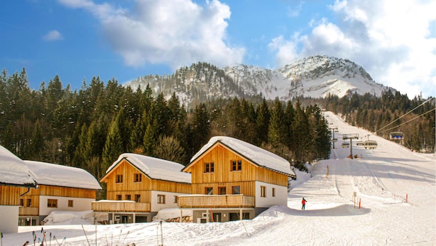 Een houten chalethuis met een met sneeuw bedekte dak, omgeven door een besneeuwd landschap en een bebost berglandschap op de achtergrond.