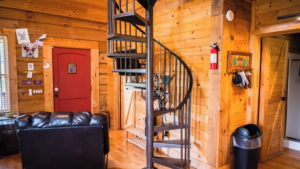 L’intérieur d’un chalet en bois, avec un escalier en colimaçon, une porte rouge, un canapé en cuir et un extincteur fixé au mur.