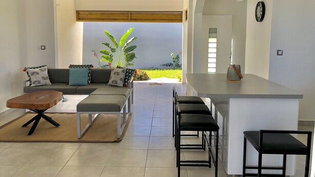 Ein modernes Wohnzimmer mit einer Couch, einem Couchtisch und Barhocker. Ein großes Fenster bietet Blick auf einen Garten.