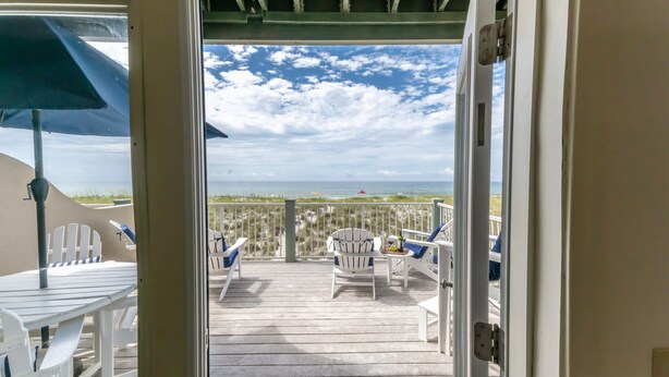Une terrasse avec des chaises blanches et une table, donnant sur une plage bordée d’une clôture et de dunes herbeuses.