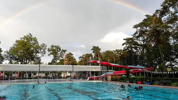Une piscine avec des personnes dans l’eau, un espace couvert avec un toit, et un toboggan aquatique rouge en arrière-plan.