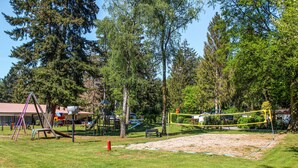 Un terrain de jeux avec des balançoires, des toboggans et un bac à sable, entouré d’arbres et avec un bâtiment en arrière-plan.