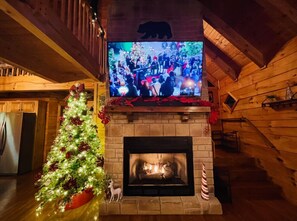 A cozy cabin interior with a fireplace, a Christmas tree, and a television displaying a festive scene.