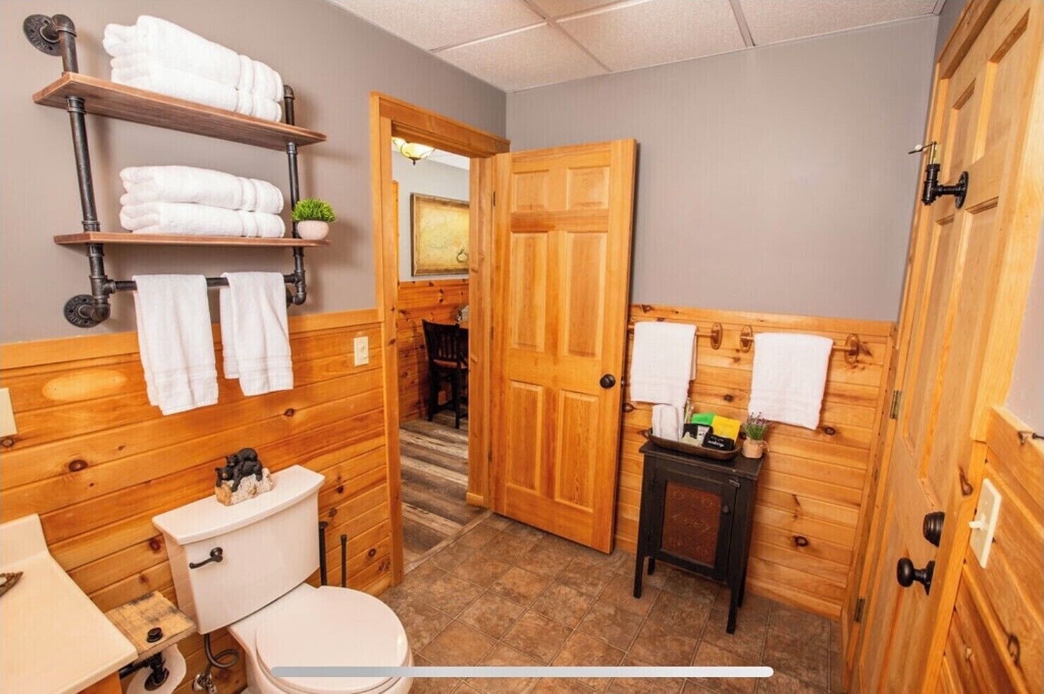 Ein Badezimmer mit Holzvertäfelung, einer Toilette, einem Waschbecken, einem Handtuchregal und einer Tür zu einem anderen Raum.