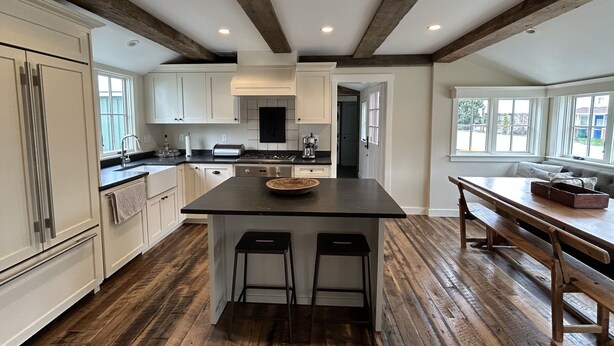 A spacious kitchen with a central island, wooden beams on the ceiling, and a view of the outdoors through large windows.