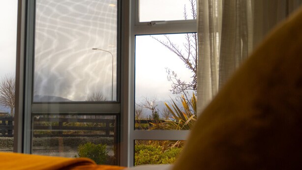 Une pièce avec une grande fenêtre, un lit recouvert d’une couverture orange, et une vue sur un jardin et des arbres à l’extérieur.