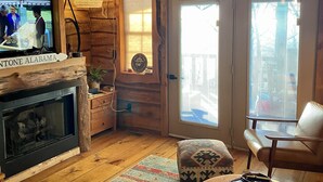 Un acogedor interior de cabaña con chimenea, paredes de madera, televisor, mesa de comedor y un ventanal grande.