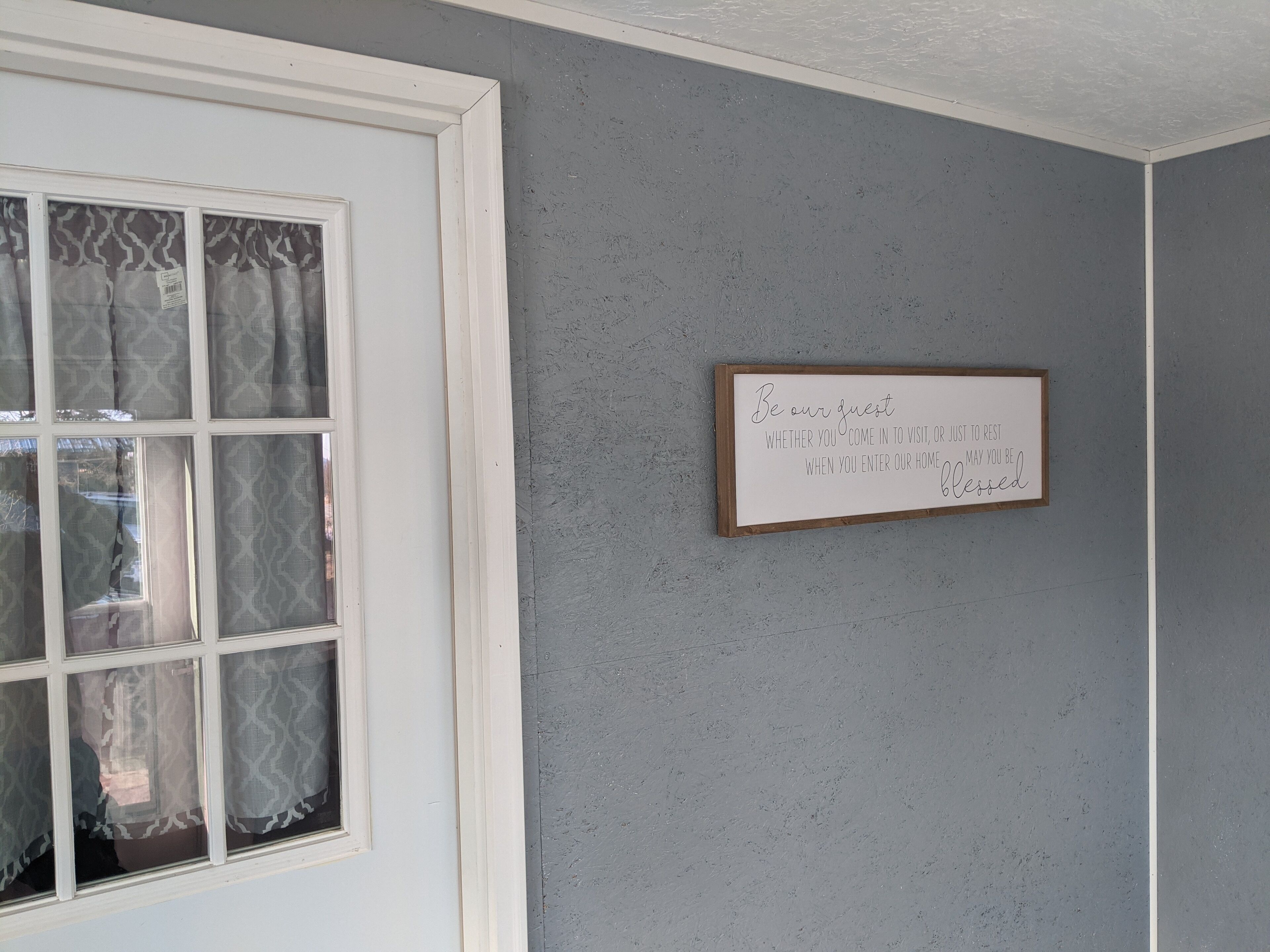 Une pièce avec un mur gris à la texture particulière, une fenêtre à cadre blanc ornée de rideaux à motifs et un panneau encadré accroché au mur.
