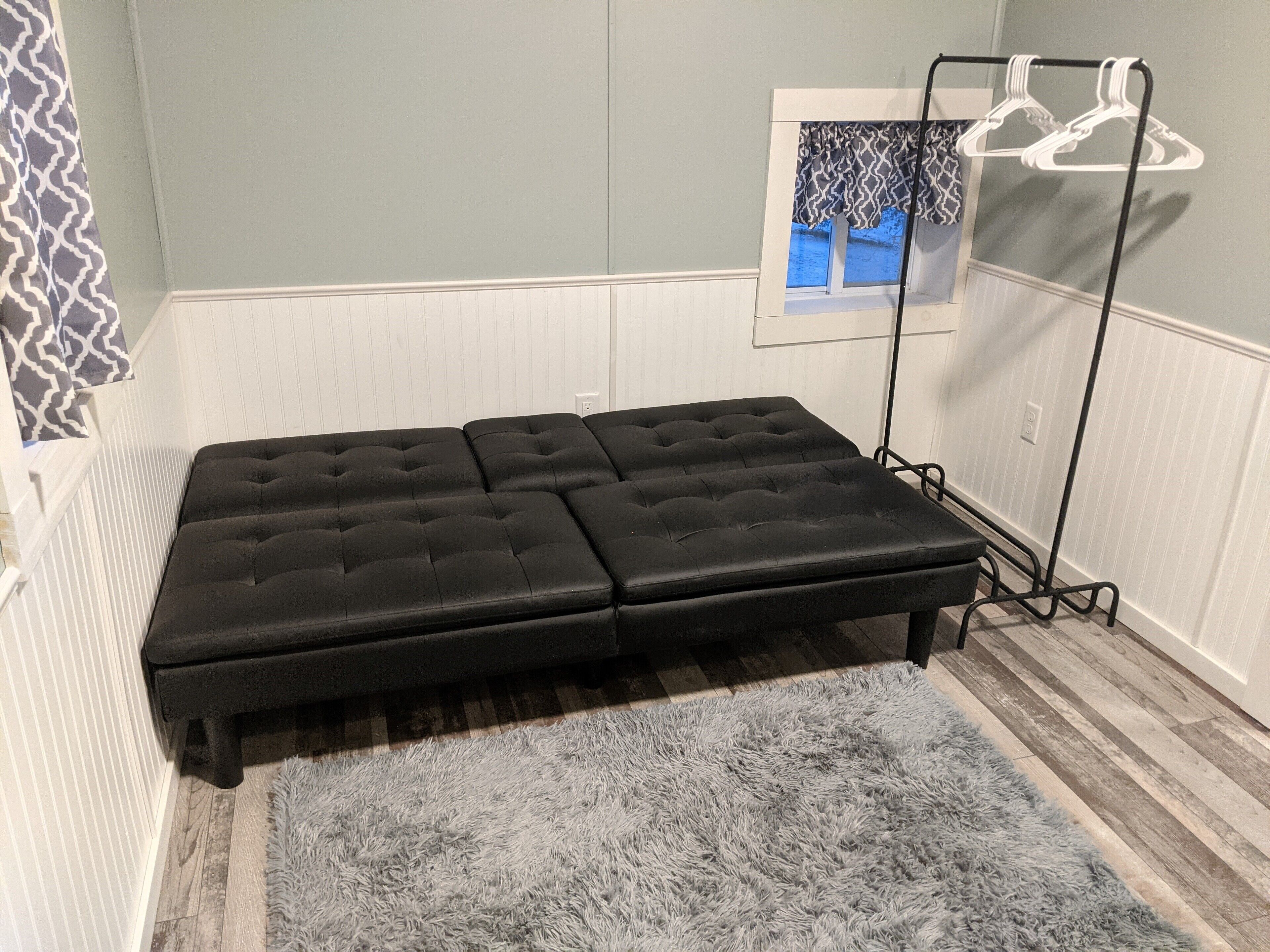 A room with a black sofa bed, a grey furry rug, a window with a view, and a clothing rack.