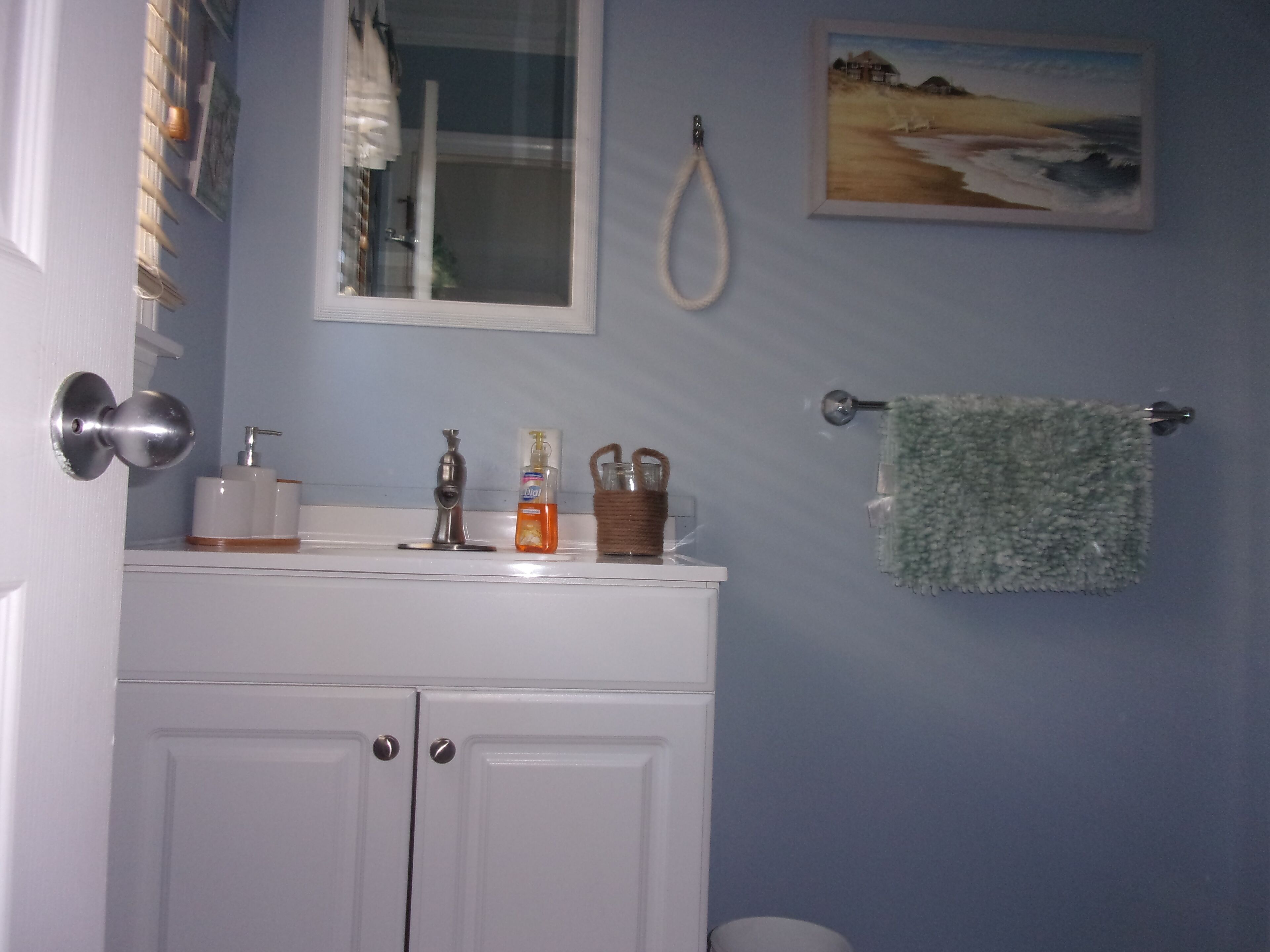 A bathroom with a white sink, a hand soap dispenser, a bottle of liquid soap, a glass, and a towel hanging on a rack.