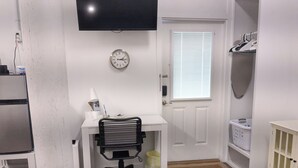 A room with a white desk, black office chair, a clock on the wall, a window with blinds, a door, and a small cabinet. - Charming studio with pier on Mobile bay (Fairhope)