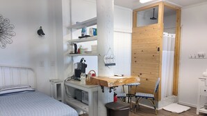A compact room with a bed, a wooden table, a coffee maker, a trash bin, and a small seating area with chairs. - Charming studio with pier on Mobile bay (Fairhope)