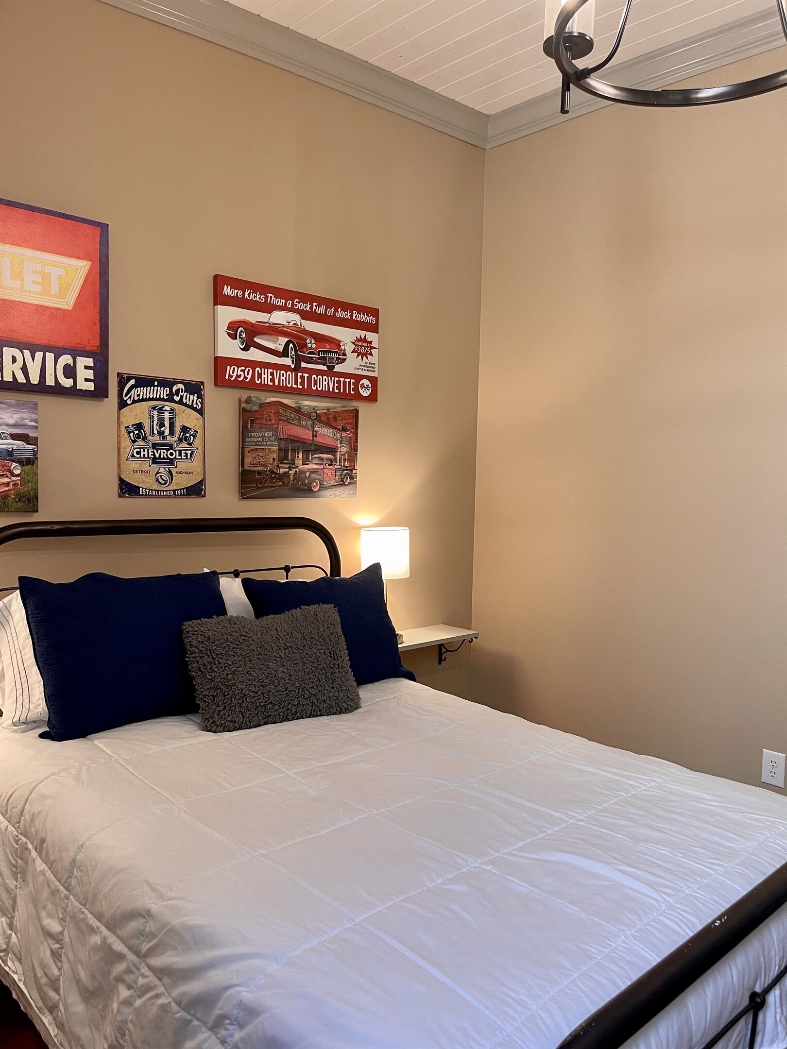 A bedroom with a bed, pillows, a nightstand, a lamp, and wall decorations including vintage automotive posters.