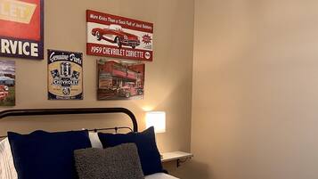 A bedroom with a bed, pillows, a nightstand, a lamp, and wall decorations including vintage automotive posters.