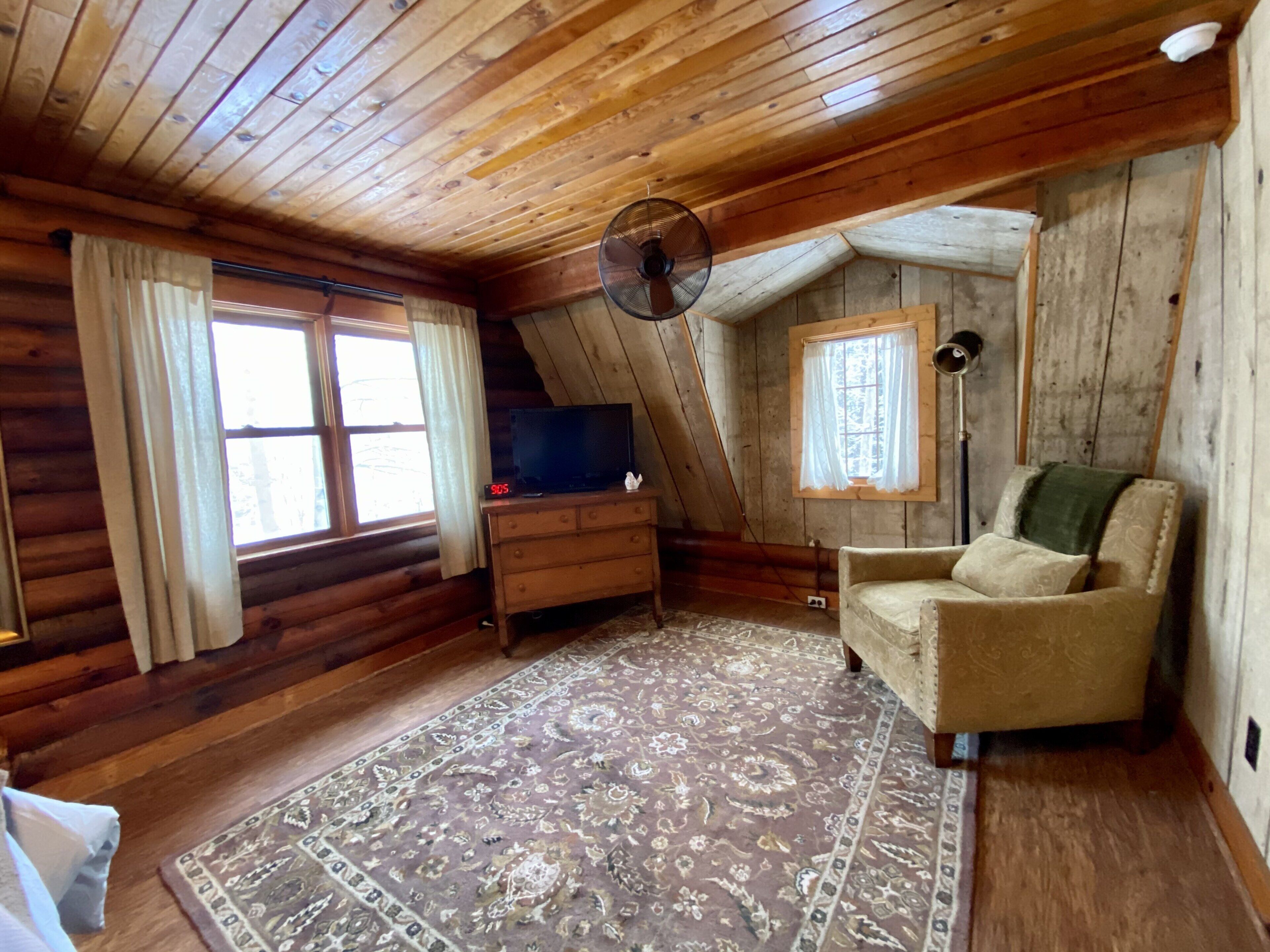 A log cabin interior with a wooden ceiling, a large rug, a sofa, a TV on a dresser, and a window with curtains.