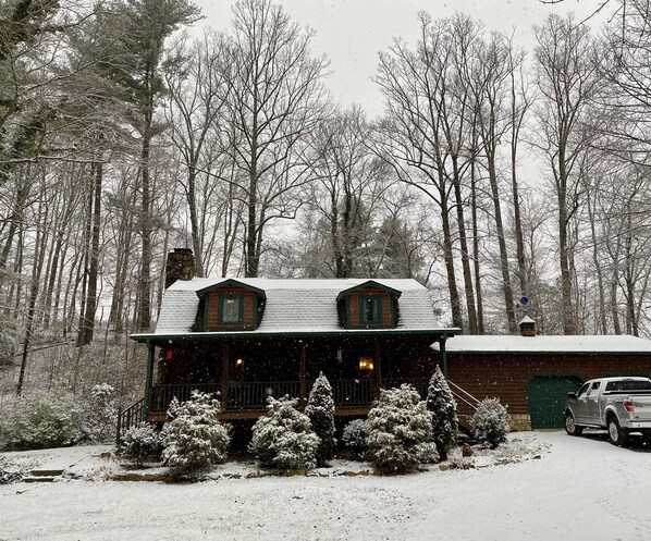 Une cabane dont le toit est recouvert de neige, entourée d’arbres et avec un véhicule garé à proximité.