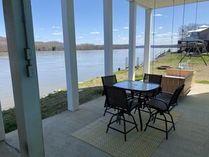 Een overdekt terras met een tafel en stoelen, uitkijkend op een meer, met een schommel en een houten terras op de achtergrond.
