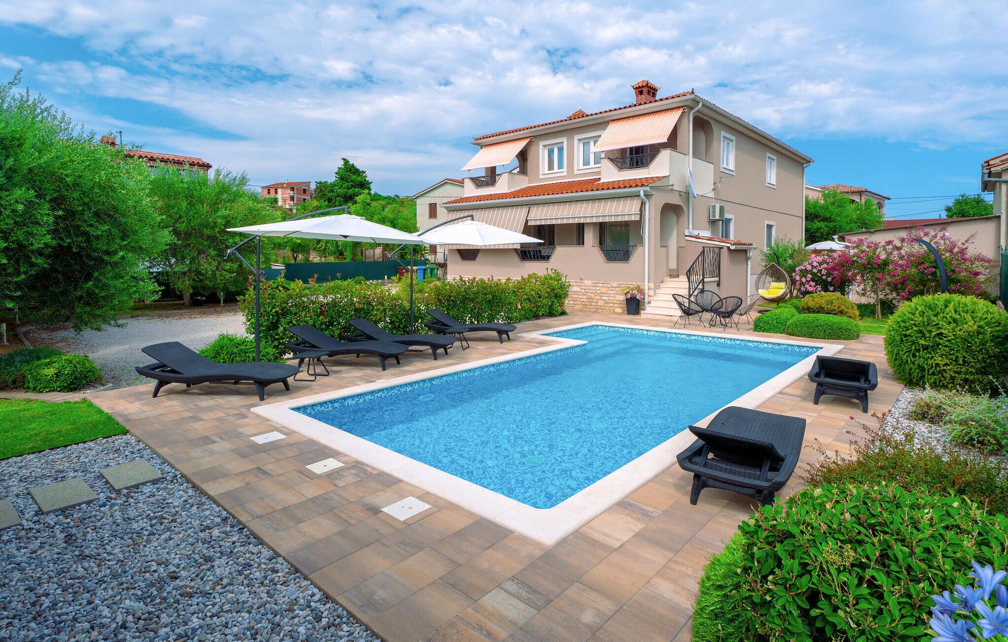 Ein Swimmingpool mit Liegestühlen, eine überdachte Sitzecke und ein zweigeschossiges Haus mit einem Ziegeldach.