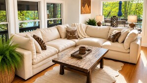A modern living room with a beige sectional sofa, a wooden coffee table, and large windows with a view of greenery. - Lakefront, 3 Amazing Decks, Tennis/Pickle Ball, Boat Slip, Hot Tub, Bed Swing
 (Oakland)